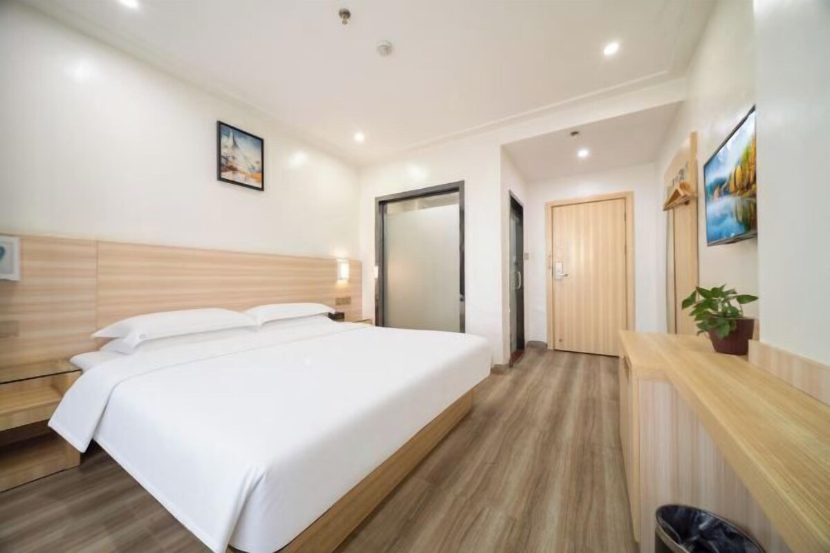 A hotel room with a large bed, wooden headboard, a bedside table, a nightstand, a wall-mounted TV, a door, and a small potted plant.