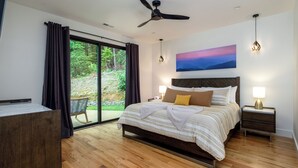 Une chambre moderne avec un grand lit, un plancher en bois, un ventilateur de plafond et une vue sur une forêt, visible à travers des portes vitrées.