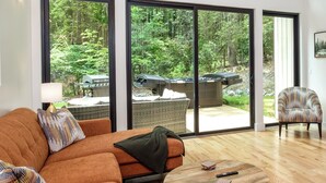 Un vaste salon agrémenté d’un grand canapé orange, d’une table basse ronde en bois et d’un fauteuil à motifs. La pièce est dotée de grandes fenêtres offrant une vue sur une forêt luxuriante et comprend un patio avec des meubles d’extérieur.