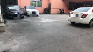 Uma área de estacionamento com três carros e duas motocicletas estacionados em frente a um edifício com fachada rosa.