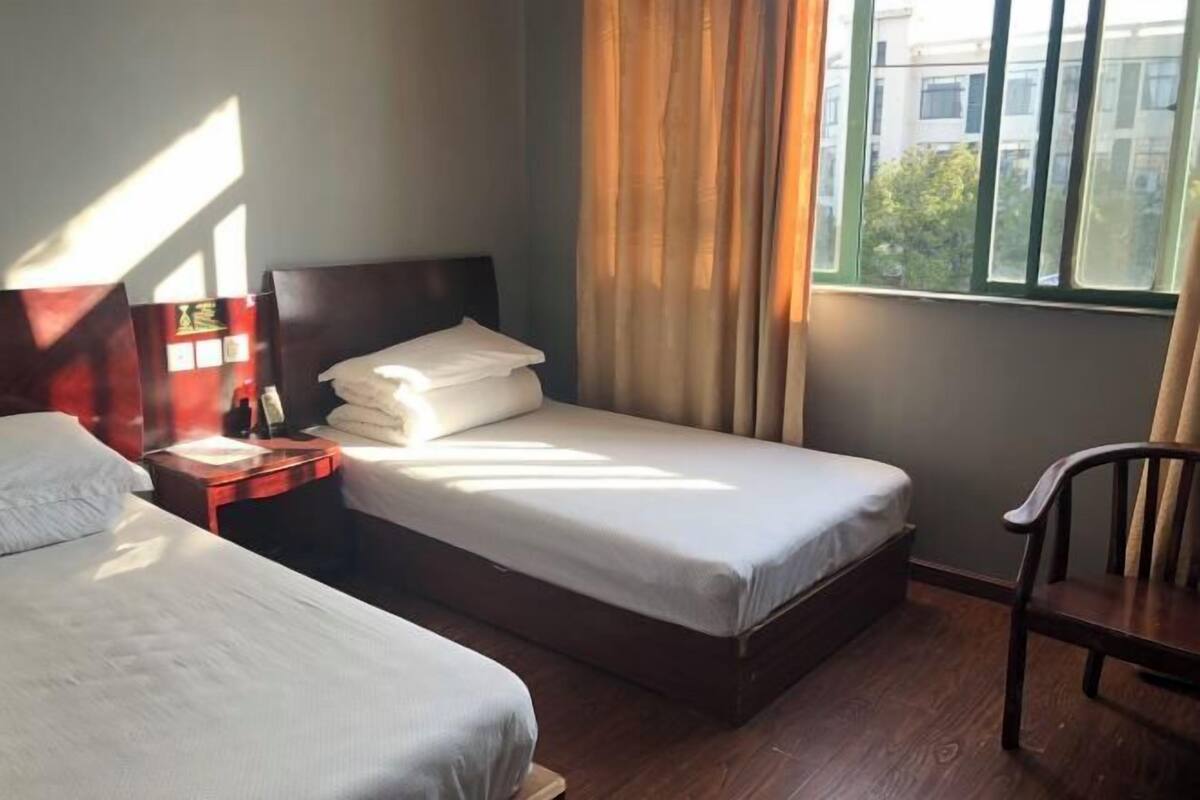 Ένα δωμάτιο ξενοδοχείου με δύο κρεβάτια, ένα κομοδίνο, μια καρέκλα και ένα παράθυρο με κουρτίνες.