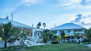 Une maison à deux étages, avec un toit bleu, entourée d’une pelouse bien entretenue et de diverses plantes tropicales.