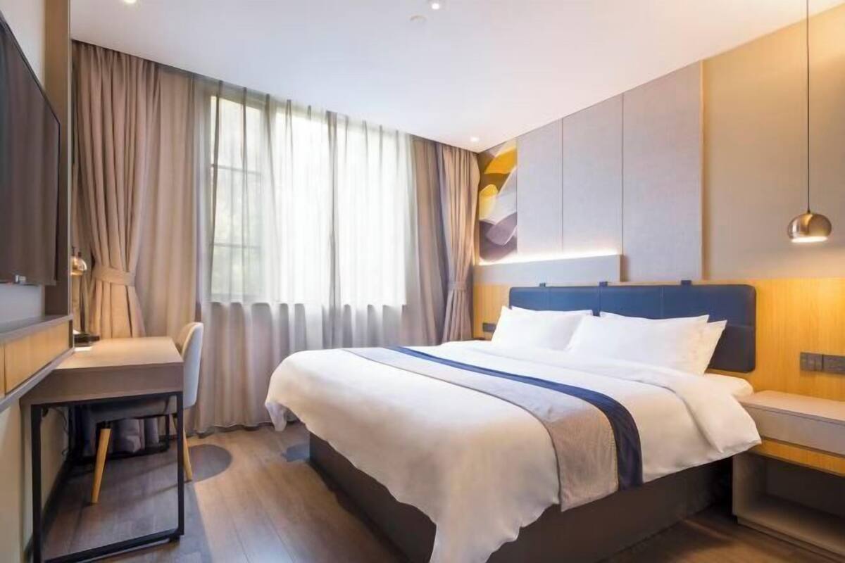 Een moderne hotelkamer met een bed, nachtkastje, bureau, stoel en een groot raam met gordijnen.