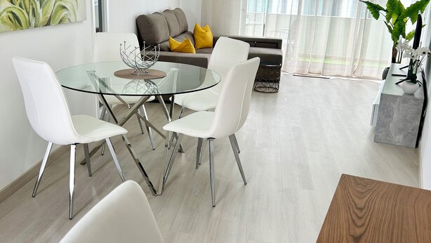 A modern living room with a glass dining table, white chairs, a grey sofa with yellow cushions, and a large window with sheer curtains. - Lido Casa by Olá Madeira (Funchal)