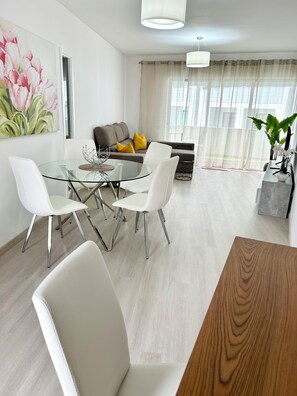 A modern living room with a glass dining table, white chairs, a grey sofa with yellow cushions, and a large window with sheer curtains. - Lido Casa by Olá Madeira (Funchal)