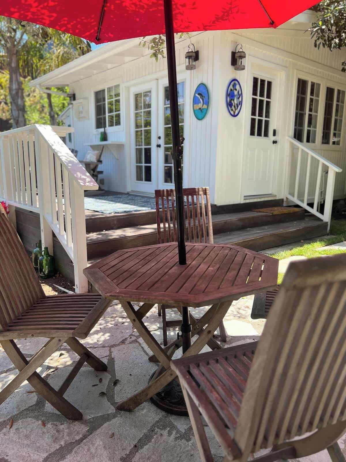 Une terrasse avec une table ronde en bois et quatre chaises, un parasol rouge et, au fond, un bâtiment blanc doté d’un porche.