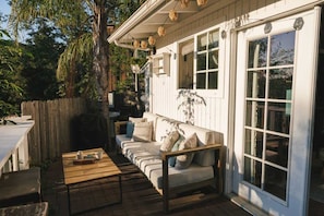 A cozy outdoor seating area with a wooden table, a cushioned sofa, and a large window with a view of the surrounding greenery. - Outdoor Clawfoot, Hot Tub, Deck (Topanga)