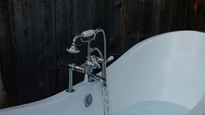 Eine freistehende Badewanne mit moderner Armatur und Duschkopf, vor einer Holzwand mit dekorativer Metallwandgestaltung.