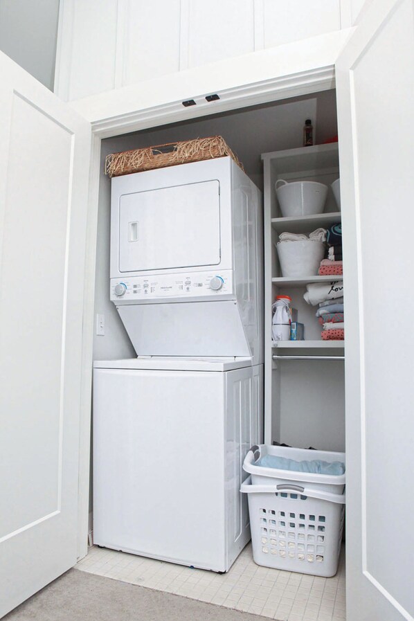 A compact laundry room with a stacked washer and dryer, a basket, and shelves with folded towels and laundry baskets.