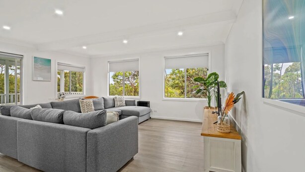 Un salón espacioso con un sofá seccional gris, ventanas amplias que ofrecen vistas a la vegetación y una mesita de madera con objetos decorativos.
