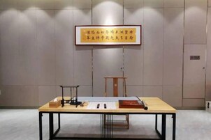 Una habitación con una mesa mostrando varios objetos, un texto enmarcado en la pared y una puerta al fondo.