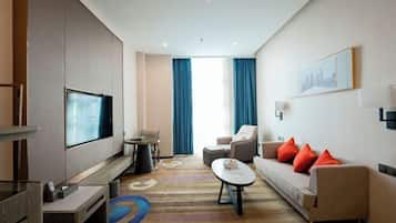A modern hotel room with a flat-screen TV, a sofa with red cushions, a coffee table, and a large window with blue curtains.