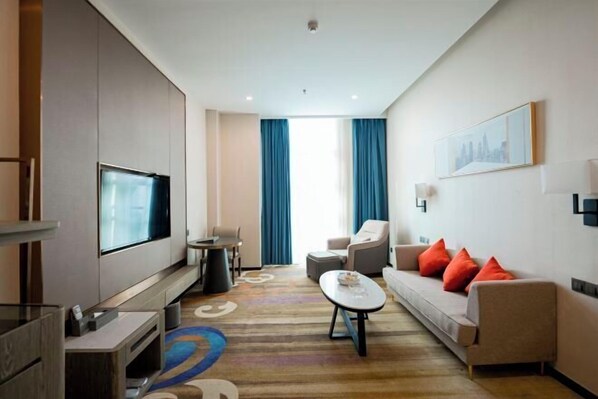 A modern hotel room with a flat-screen TV, a sofa with red cushions, a coffee table, and a large window with blue curtains. - Haomei International Hotel (Anxi Government Store) (Anxi)