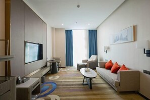 A modern hotel room with a flat-screen TV, a sofa with orange cushions, a coffee table, a desk with a chair, and a large window with blue curtains. - Haomei International Hotel (Anxi Government Store) (Anxi)