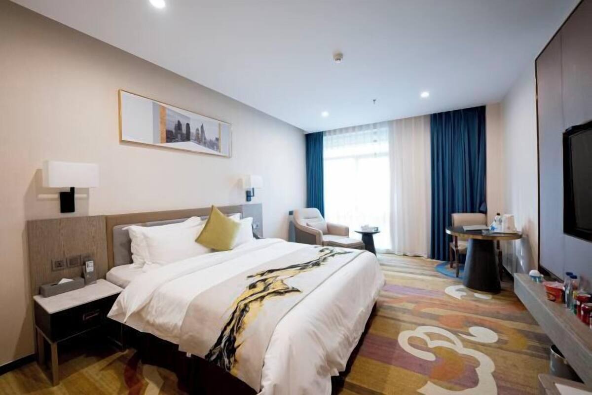 A hotel room with a bed, bedside tables, a chair, a TV, and a window with curtains.