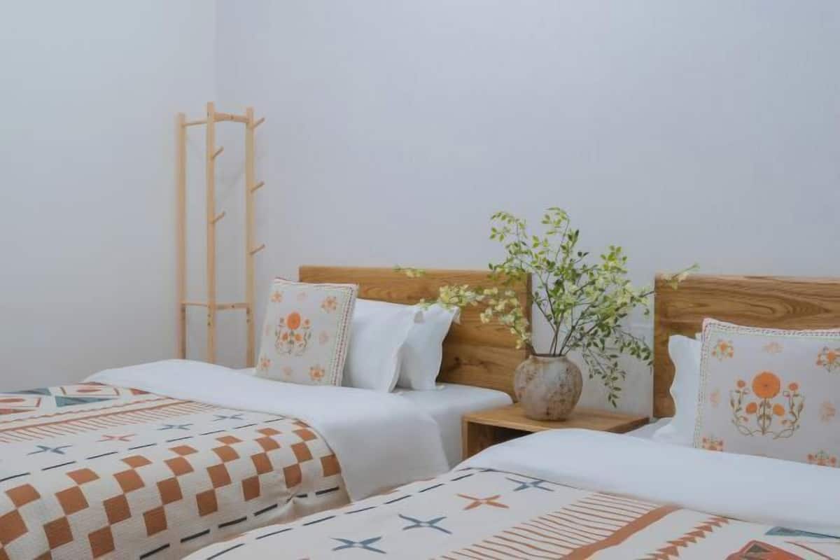 Une chambre avec deux lits, une tête de lit en bois, une table de chevet ornée d’un vase de fleurs et une échelle en bois appuyée contre le mur.