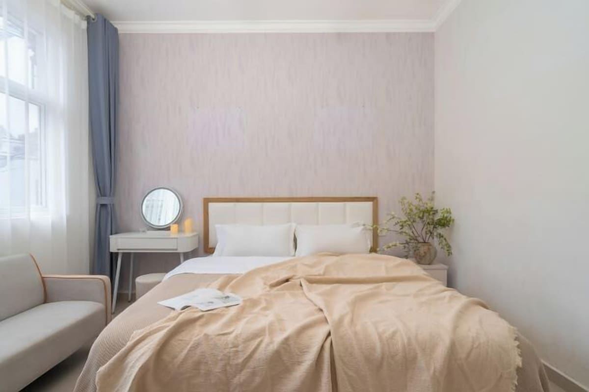 A hotel room with a bed, a bedside table with a lamp, a small round mirror, a potted plant, and a window with curtains.
