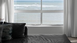 Una habitación con un gran ventanal con vistas al océano, una cama con colchón gris y cortinas blancas.