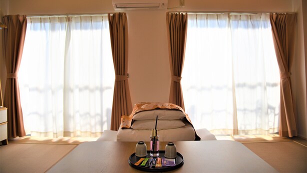 Une chambre d’hôtel avec un lit, une table sur laquelle se trouve un plateau avec des articles de toilette, et de grandes fenêtres avec des rideaux.