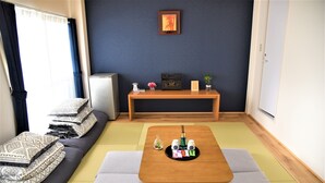 Une pièce avec un sol recouvert de tatamis, une petite table basse en bois avec un service à thé, un canapé avec des coussins et un petit réfrigérateur.