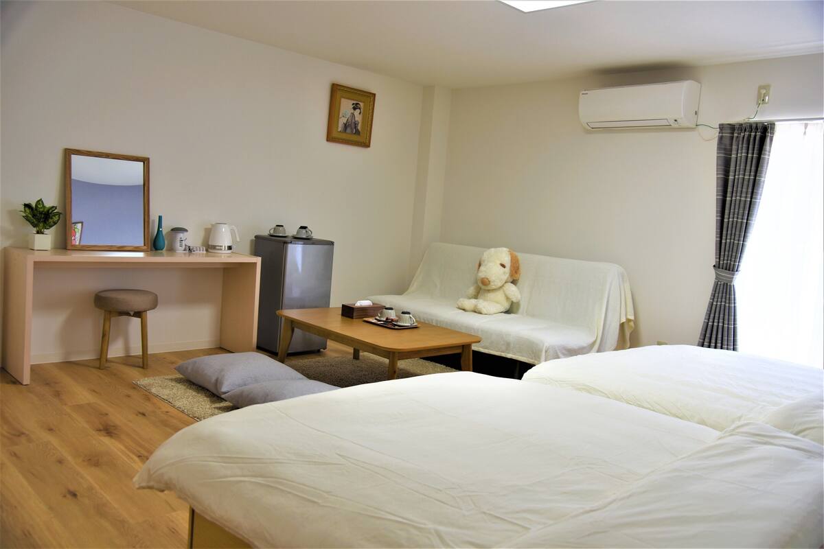 Hotelkamer met twee bedden, een sofa, een bureau met stoel, een koffiezetapparaat, een waterkoker, een kleine plant en een airconditioning.