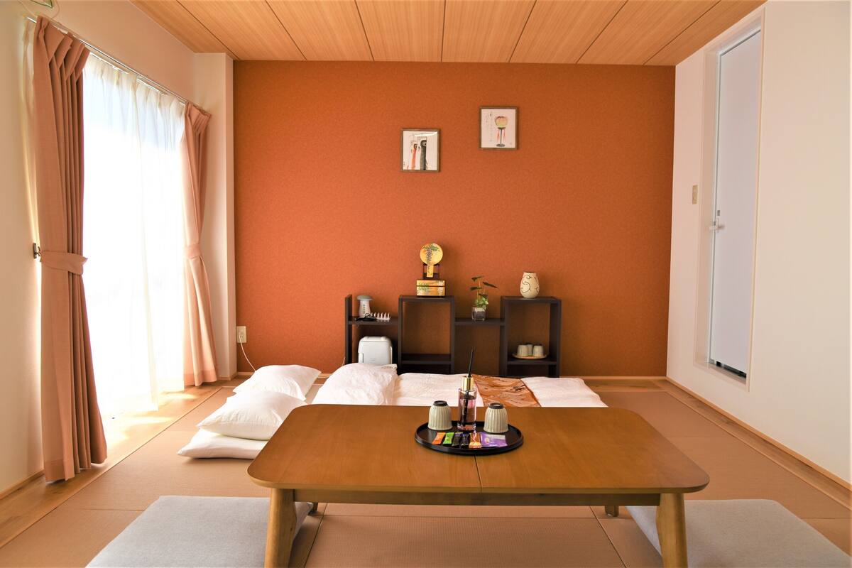 Een kamer met een houten tafel, een bed met witte lakens, een klein plankje met decoratieve spullen, en een raam met gordijnen.