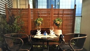 Une table ronde avec deux chaises, un verre d’eau et une théière, le tout placé sur un balcon doté d’une balustrade en bois et de plantes en pot.