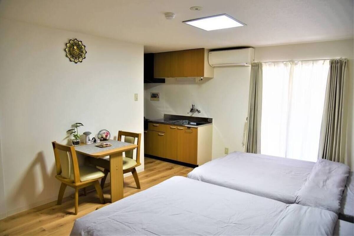 Een kamer met een bed, een bureau met stoel, een kleine kitchenette en een raam met gordijnen.