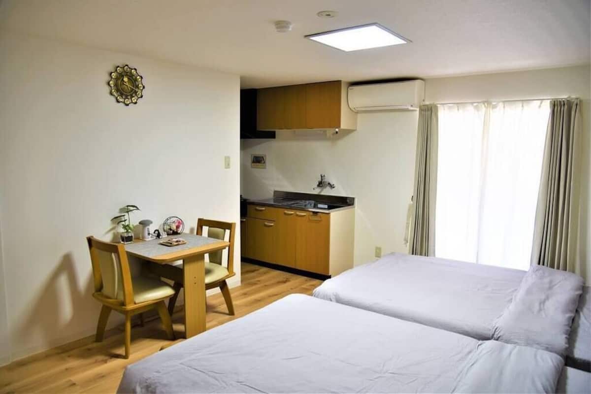 Una habitación con cama, escritorio y silla, una pequeña kitchenette y ventana con cortinas.