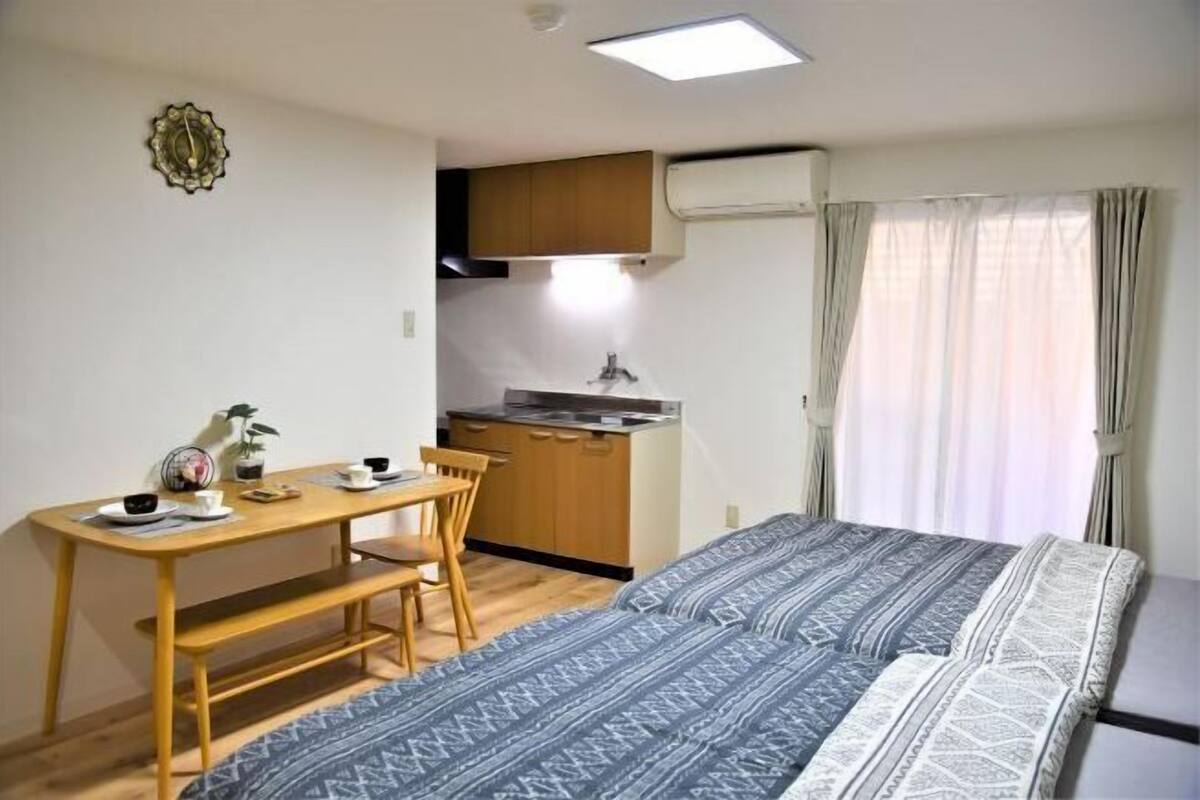 Een compacte kamer met een bed, een kitchenette, een eettafel met stoelen en een raam met gordijnen.