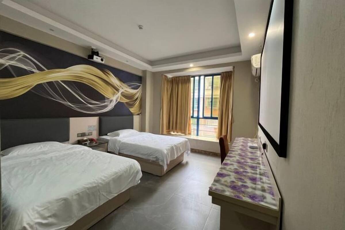 A hotel room with two beds, a desk, a large window with curtains, and a wall-mounted TV.