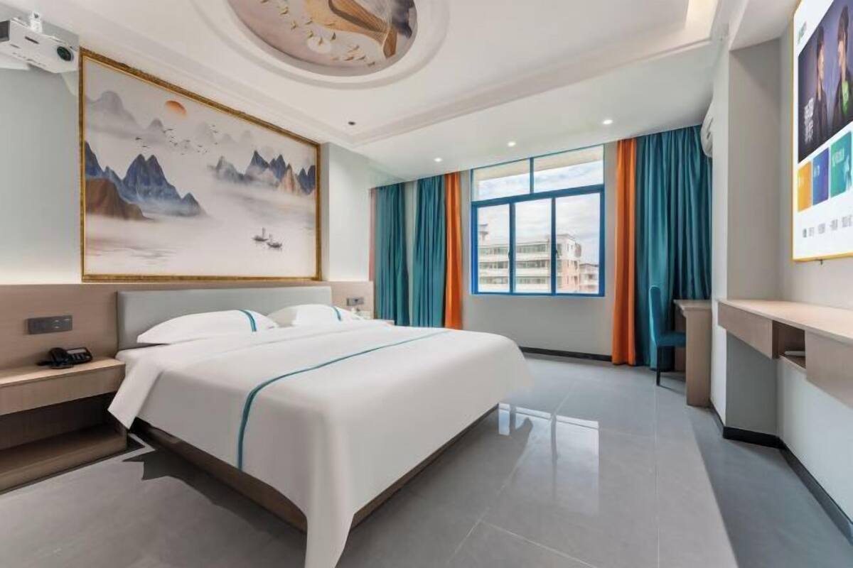 A hotel room with a large bed, a desk, a chair, a painting on the wall, and a window with curtains.