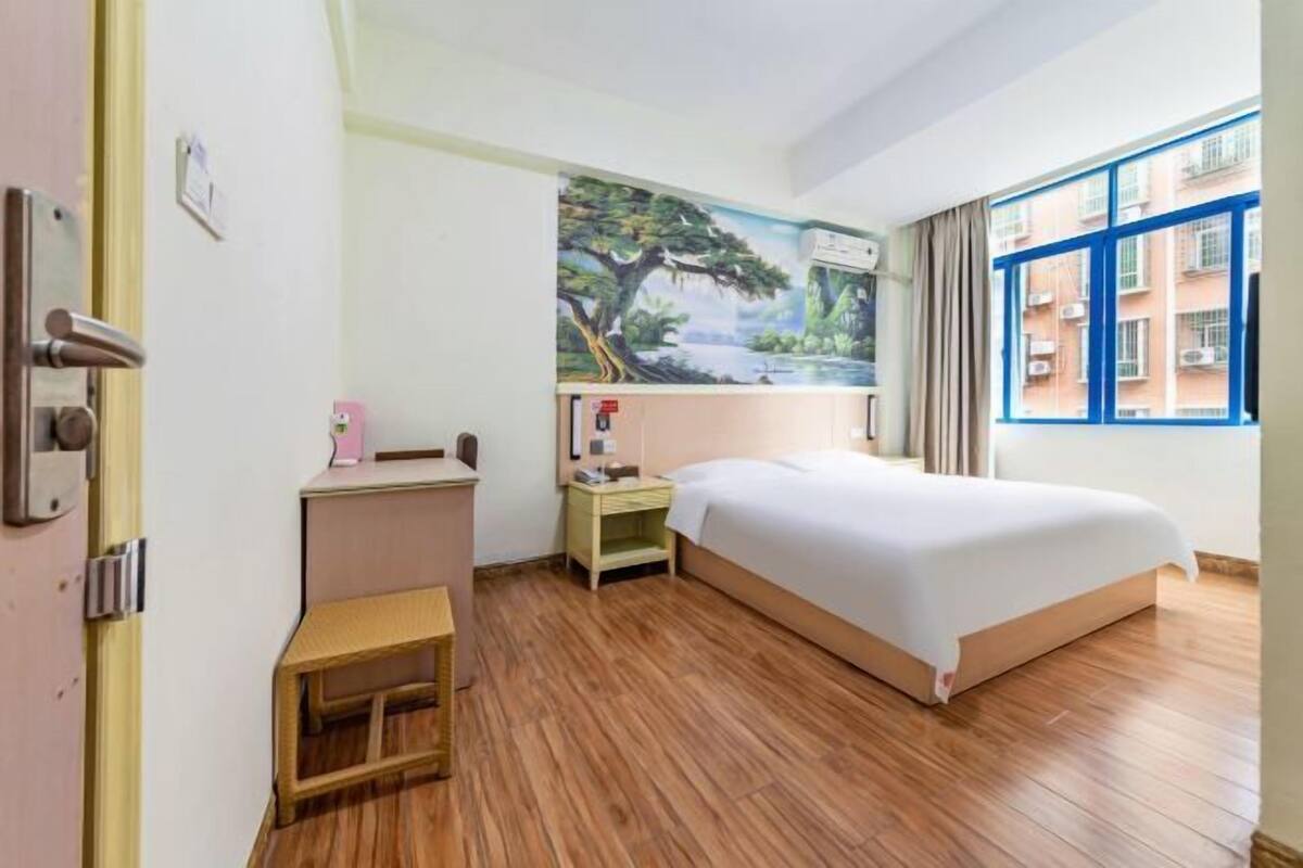 A hotel room with a bed, a nightstand, a small table, a painting on the wall, and a window with a view of buildings.