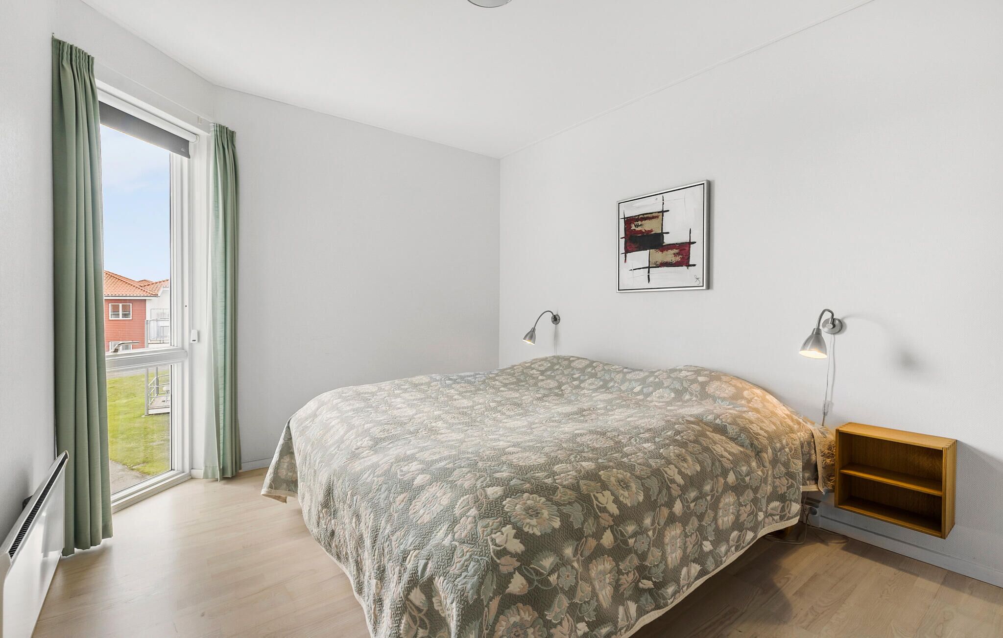 A bedroom with a large bed, a window with curtains, a wall-mounted shelf, and a framed picture.