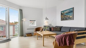 A modern living room with a sofa, a dining table, and chairs. There is a large window with a view of buildings outside. - Gudhjem Søpark Lejl. 18 (Gudhjem)