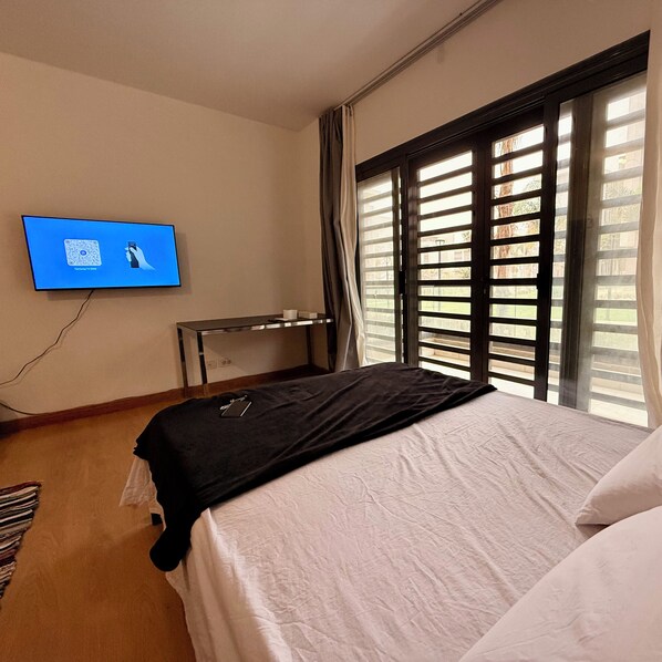 A hotel room with a bed, a desk, a TV mounted on the wall, and a window with blinds.