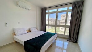 A bedroom with a bed, an air conditioner, a window with curtains, and a view of buildings outside. - Madinaty (New Cairo)