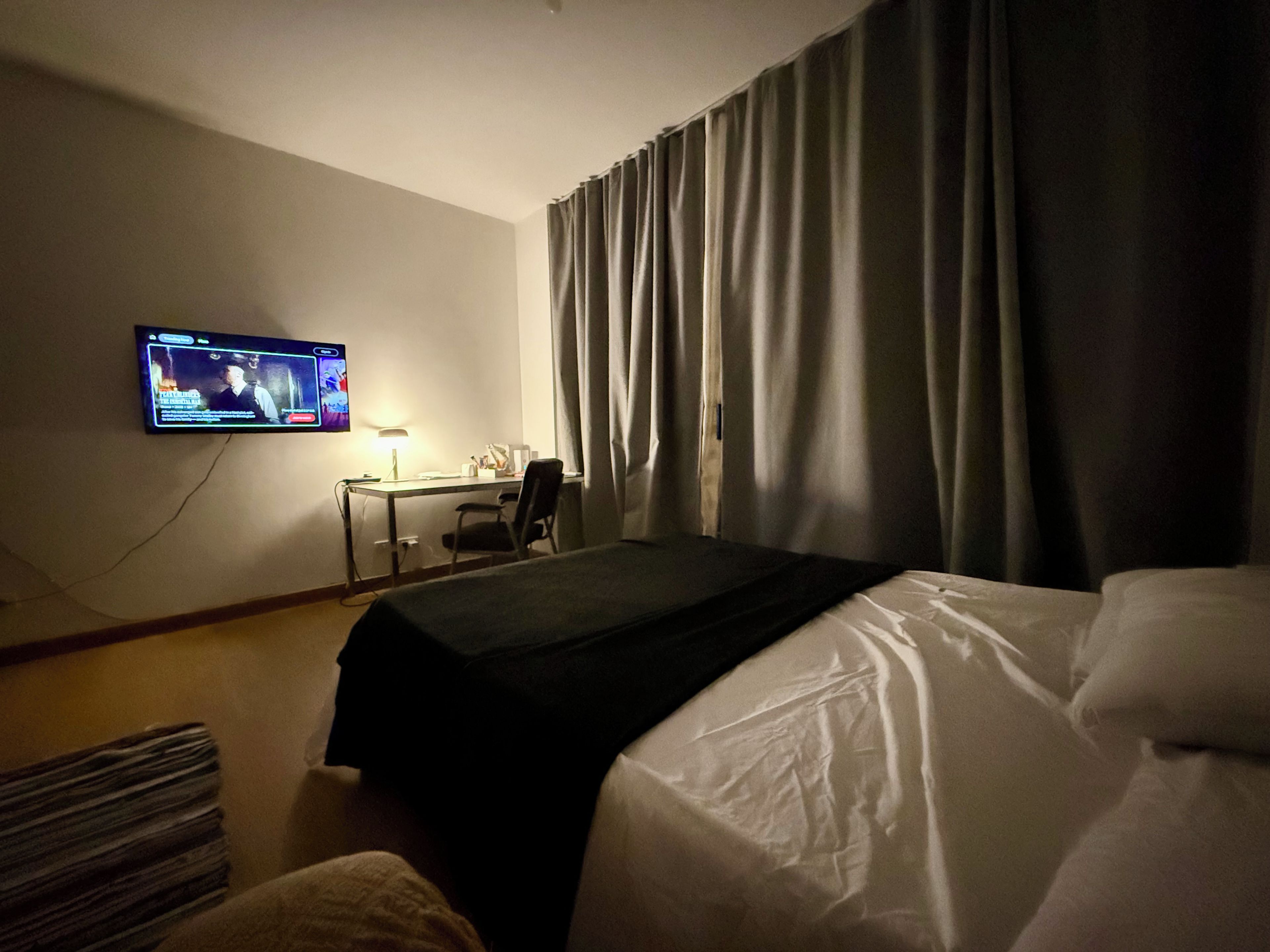 A hotel room with a bed, a desk, a chair, a television mounted on the wall, and a window with curtains.