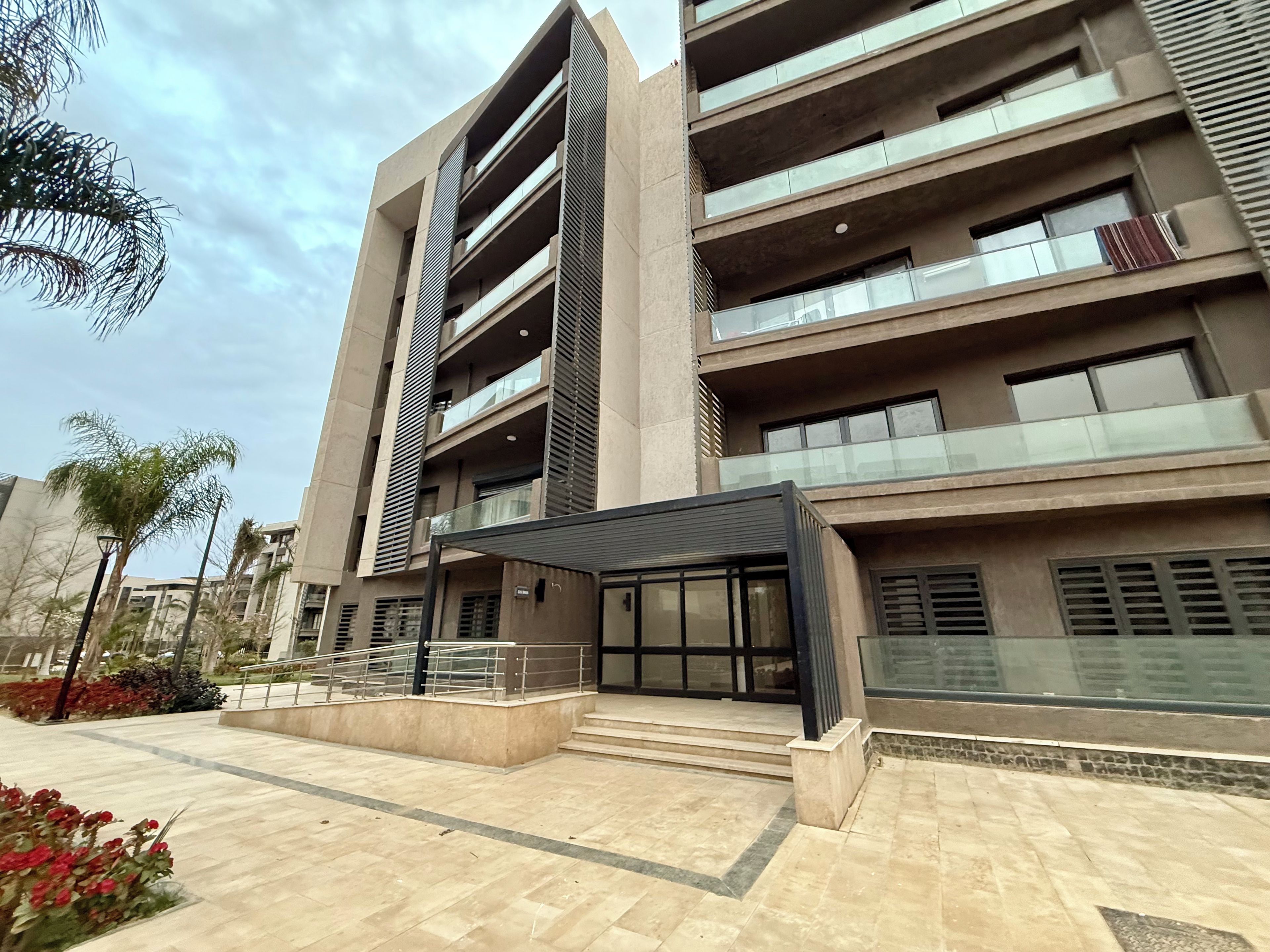 A modern multi-story building with balconies, a covered entrance, and a landscaped area with plants.