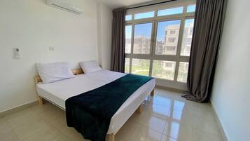 A bedroom with a bed, an air conditioner, a window with curtains, and a view of buildings outside.