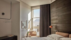 Une chambre moderne avec une grande fenêtre, une tête de lit en bois, une table de chevet avec une lampe et un lit recouvert de draps blancs.