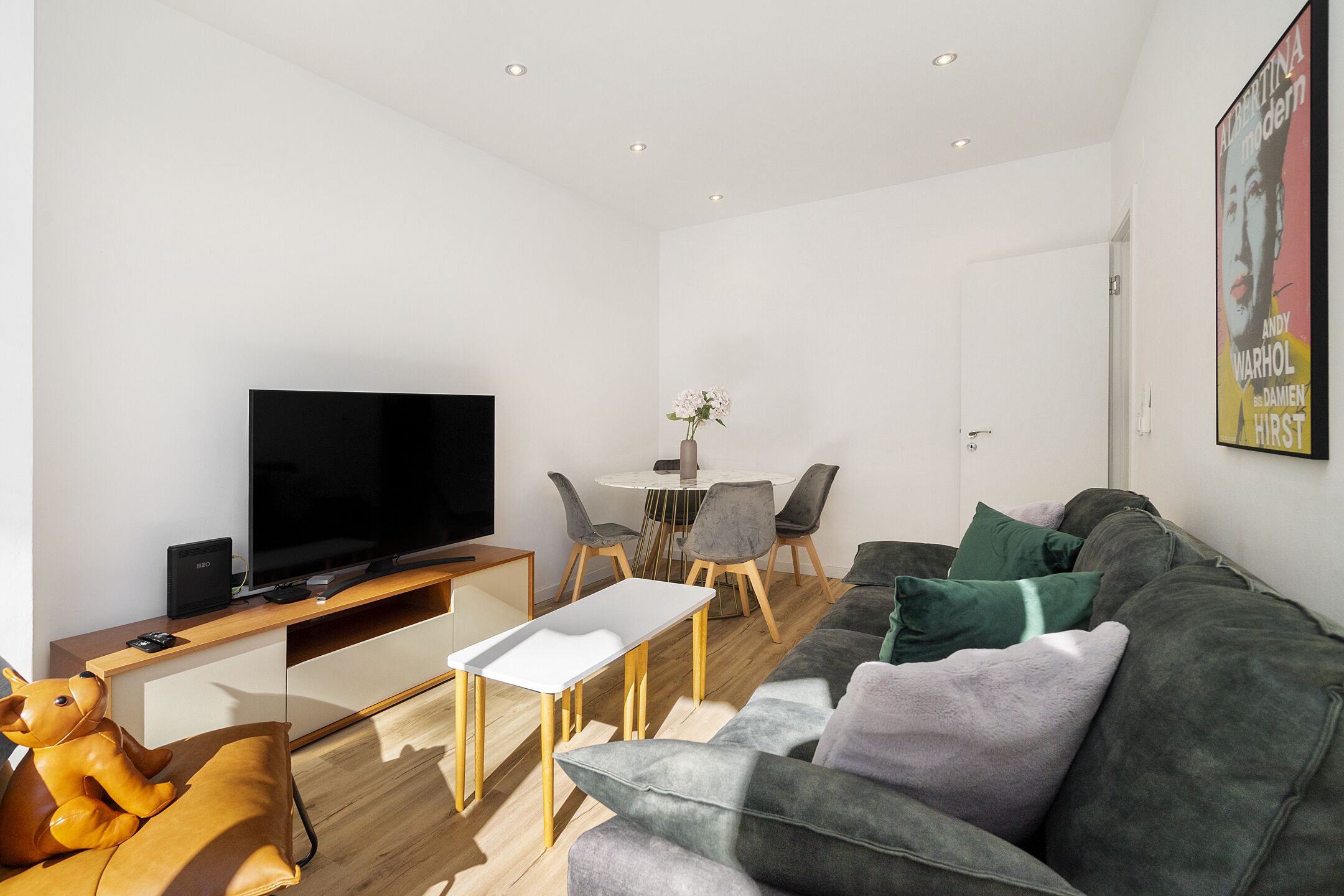A modern living room with a flat-screen TV, a sofa, a coffee table, and a dining area with chairs.