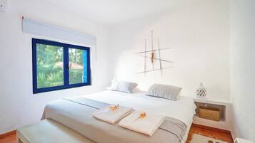 Un dormitorio con una cama grande, una ventana con vistas a zonas verdes, una lámpara de pared y una mesita de noche con una lámpara y una cesta.