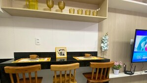 A modern kitchen with a black countertop, three wooden bar stools, a shelf with decorative items, and a TV on the wall. - Flat Ofurô 06 VP by La Playa (Ipojuca)