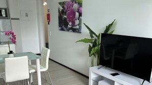 A modern living room with a flat-screen TV, a glass dining table with chairs, a coffee table, and a large potted plant.