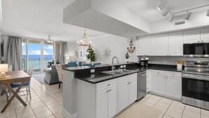 A modern kitchen with white cabinets, a black countertop, and stainless steel appliances. There is a dining area with a table and chairs, and a view of the ocean through large windows. - Gulf-Front 2 Bed, 2 Bath • Sleeps 8 • Amenity-Packed • Beach Chair Service for 2 (Panama City Beach)