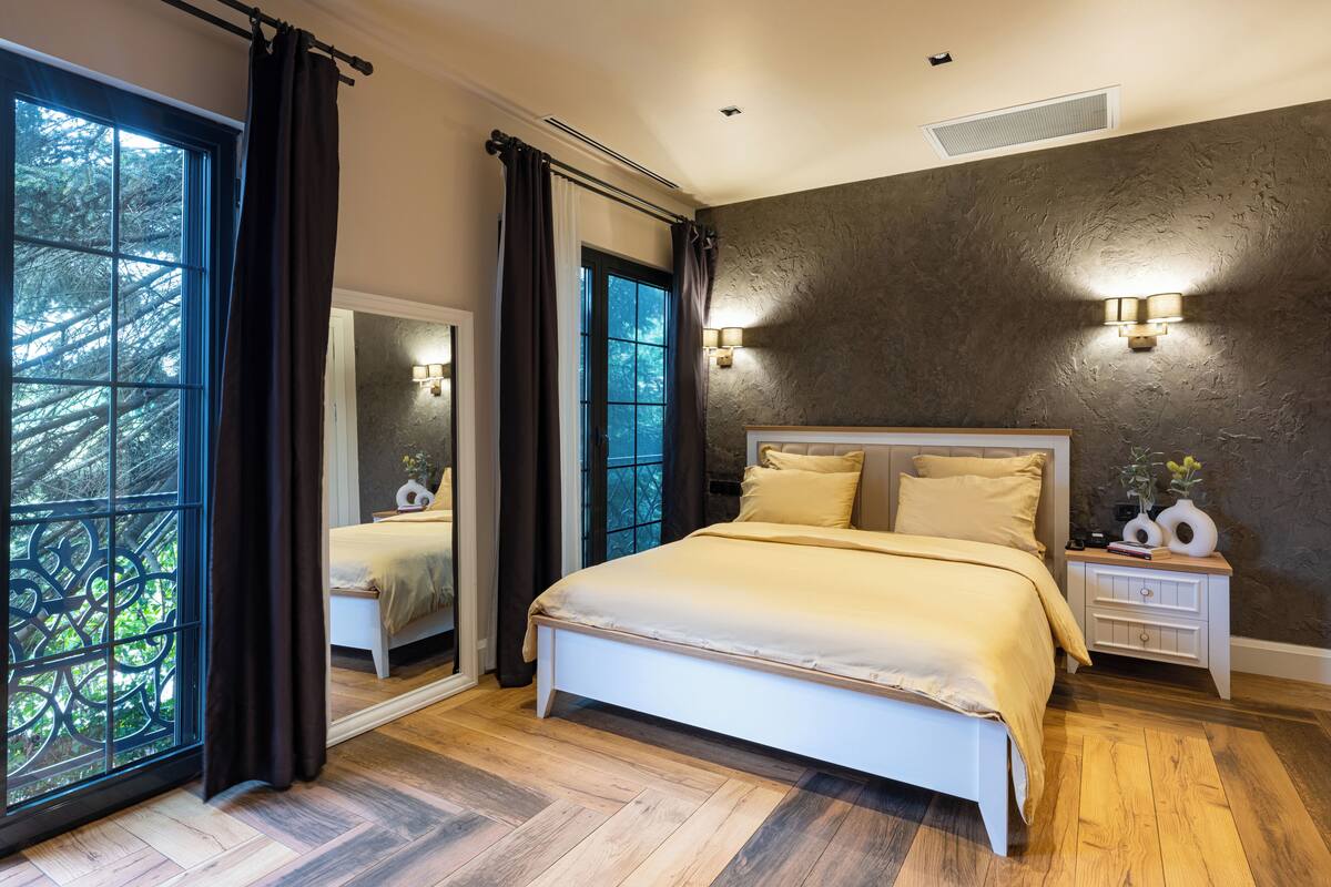 A bedroom with a large bed, a nightstand, a mirror, and a view of trees through a window.