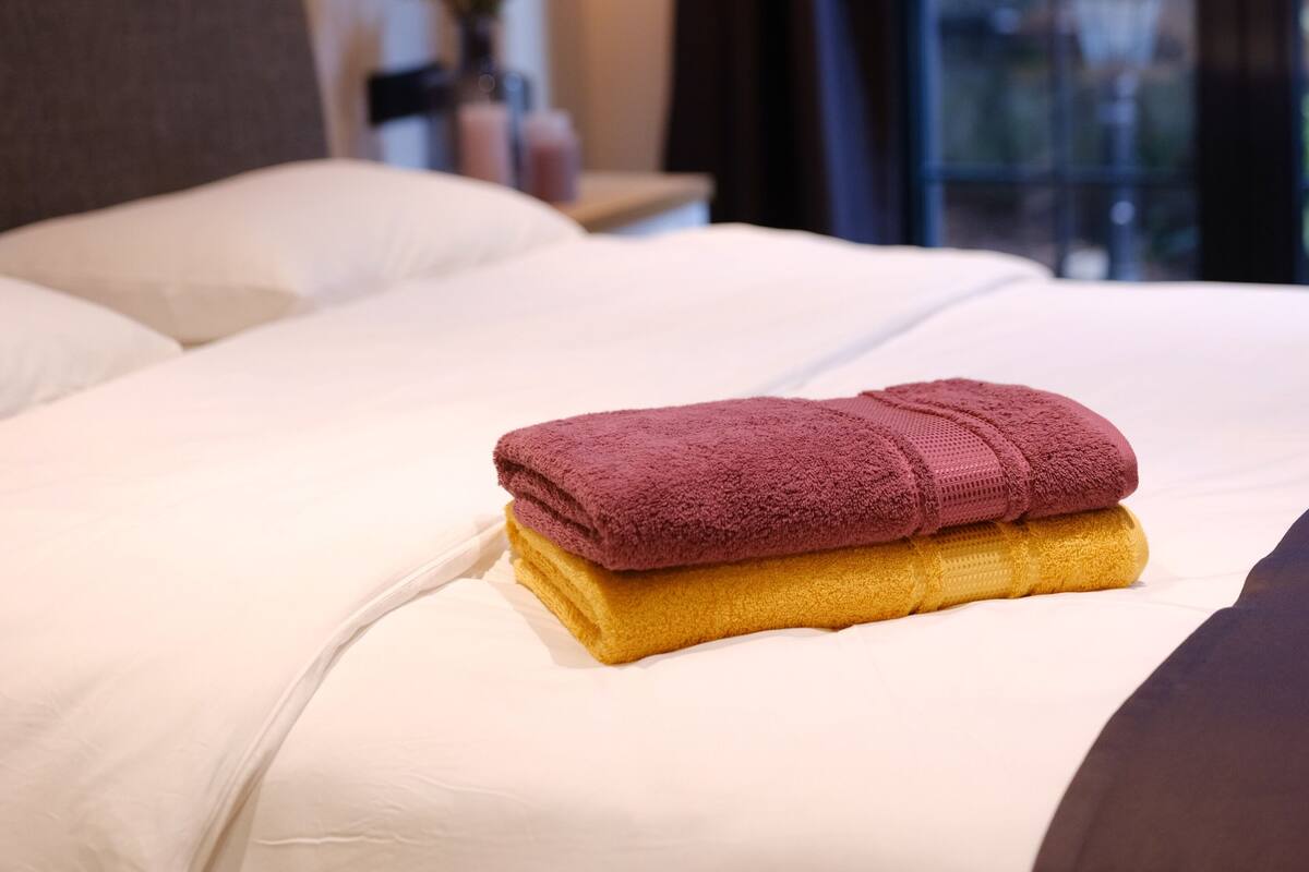 A neatly made bed with a purple and yellow towel, a lamp, and a window with a view of greenery.