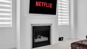 A modern living room with a fireplace, a mounted television displaying the Netflix logo, and large windows with blinds. - Beautiful New Build by Elora (Fergus)