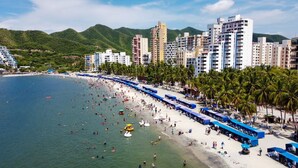 Una playa con muchas personas, una fila de cabañas azules frente al mar y un telón de edificios y colinas.
